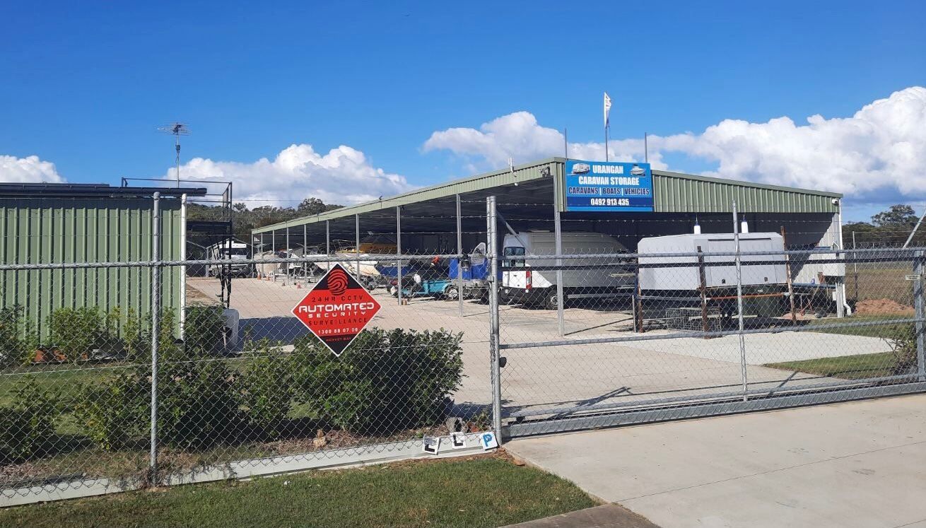 The gated entrance to Urangan Caravan Storage, with a long green Colorbond covered shelter and caravans parked on the concrete hardstand under a clear blue sky.
